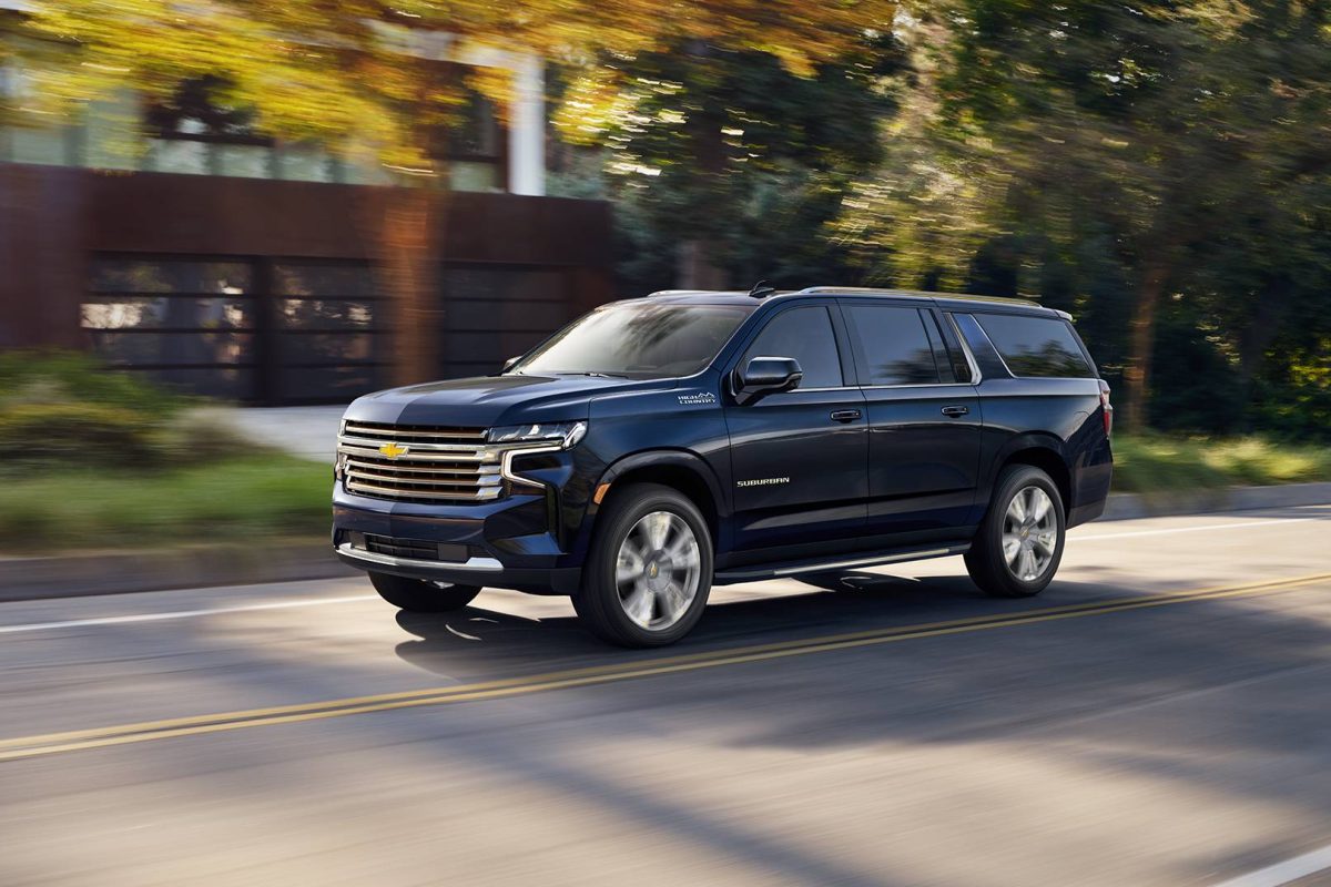 A dark blue Chevrolet Suburban SUV, often used for airports transportation, drives down a tree-lined street with a modern building in the background, captured in motion on a sunny day.