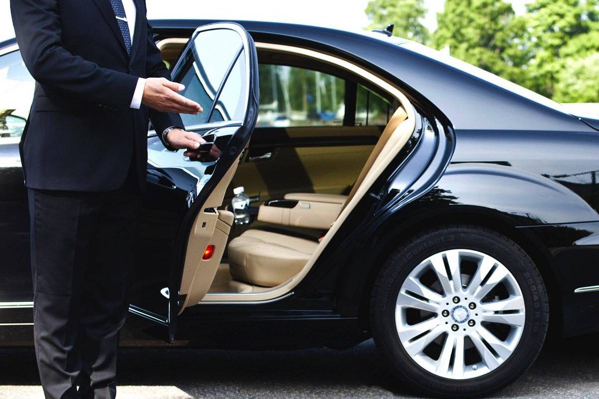 A chauffeur in a suit holds open the rear door of a black luxury car, ready for a passenger arriving for Airports Transportation. The car is parked outdoors on a sunny day with green trees in the background.