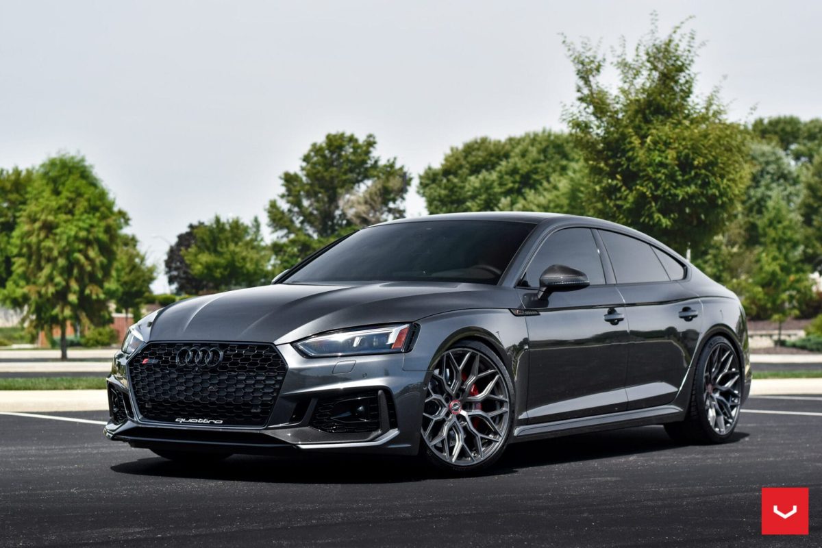 A sleek gray Audi RS5 sedan with tinted windows, perfect for airports transportation, is parked on an asphalt lot surrounded by green trees and grass on a sunny day. The car features sporty rims and a prominent front grille.