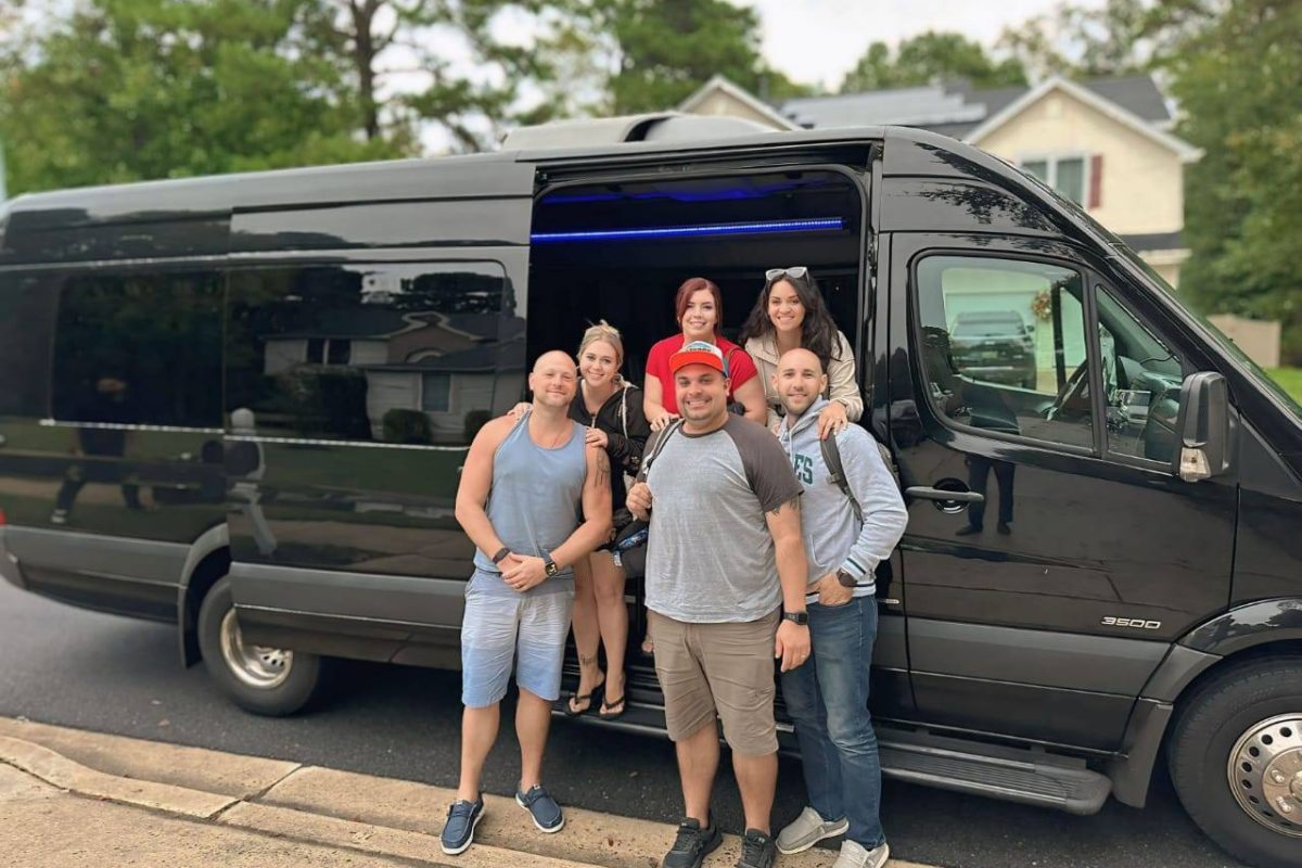 Six adults pose and smile in front of a black Airports Transportation van parked on a suburban street. Three people stand on the sidewalk, while three stand inside the van by the open door. Houses and trees are visible in the background.