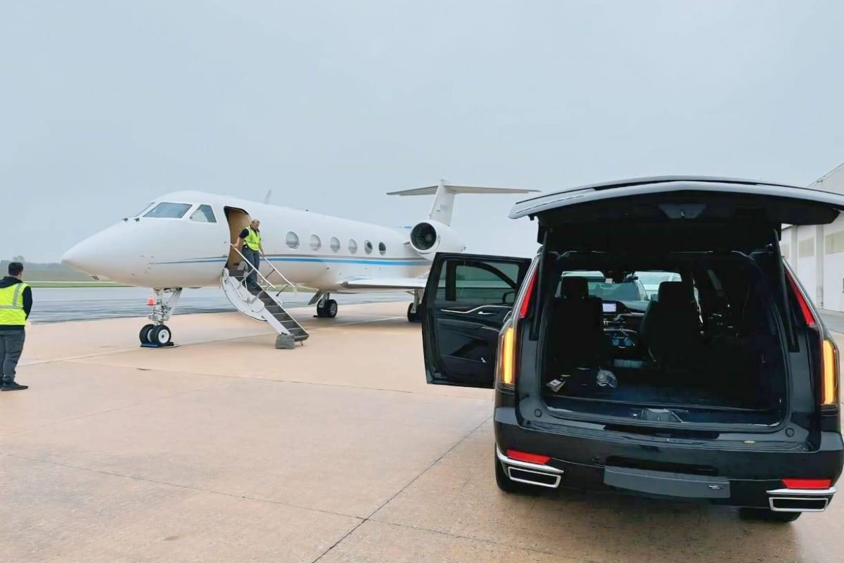A private jet is parked on the runway at one of the bustling airports, with a person on the stairs and another nearby. In the foreground, an SUV used for transportation has its trunk and door open beside the plane.